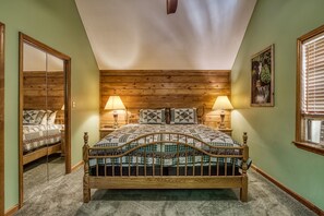 A bedroom with a wooden headboard, two bedside lamps, a bed with patterned bedding, a mirror, and a window with blinds.