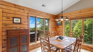 Interior de una cabaña de madera con una mesa de comedor, sillas y ventanas amplias que ofrecen vistas al exterior.