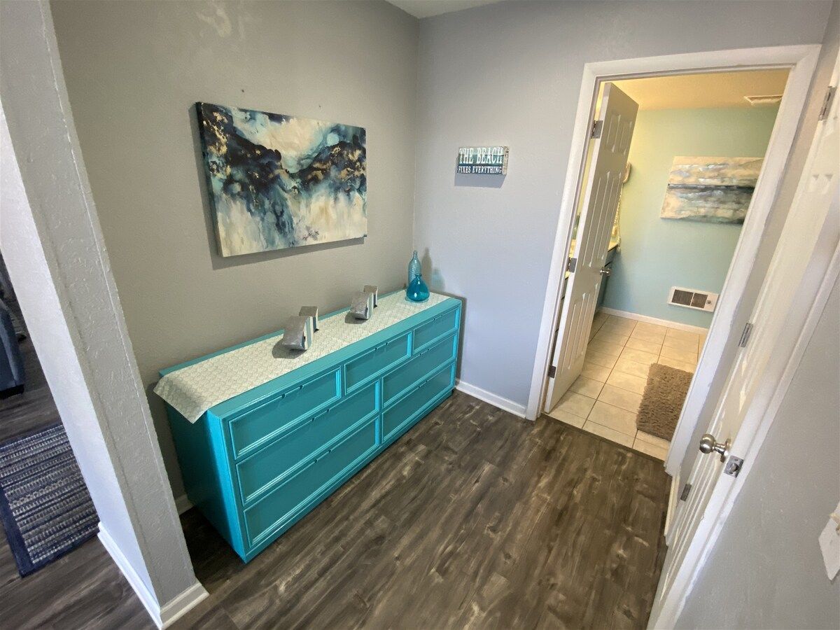 A hallway with a teal vanity, a painting on the wall, and a door leading to another room.