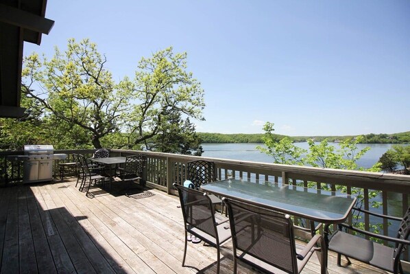 A wooden deck with a grill, table, and chairs overlooking a lake.