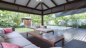 Un espacio exterior techado con terraza de madera, piscina y zona de estar con sofá y mesa.