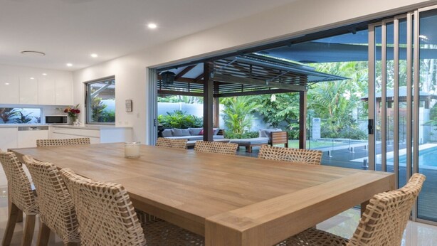Una cocina moderna con armarios blancos, un comedor con mesa de madera y sillas de mimbre, y un área de estar al aire libre con vistas a la vegetación.