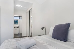 Hotellihuone, jossa on sänky, yöpöytälamppu, rullalle taitettu pyyhe ja näkymä kylpyhuoneeseen.
