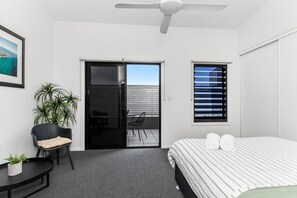 Une chambre d’hôtel avec un lit, une chaise, une petite table, une plante en pot, une porte coulissante et une fenêtre avec des stores.
