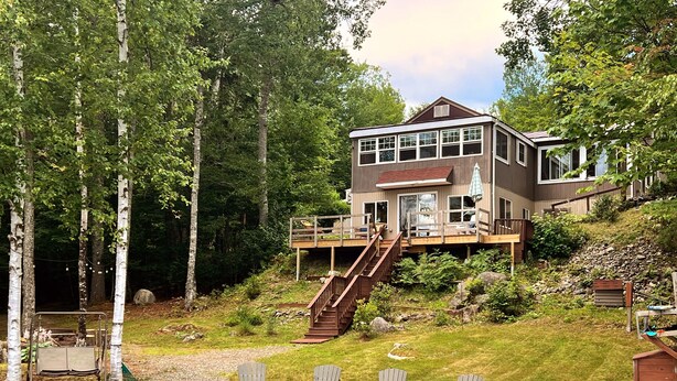 Una casa de dos pisos con un balcón y escaleras que conducen a un segundo nivel, rodeada por un bosque y un lago.