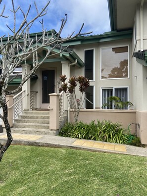 Un edificio de dos pisos con techo verde, una entrada frontal con escaleras y un pequeño jardín con plantas y un árbol.