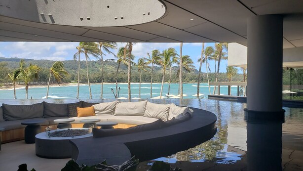 A modern interior with a curved sofa, round tables, and a view of a beach with palm trees. -  Location! Golf, Swim, Surf & WALKING distance to Hotel and beaches. (Kahuku)