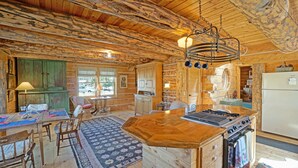 A rustic cabin interior with a wooden dining table, chairs, a kitchen area with a stove, and a large window.