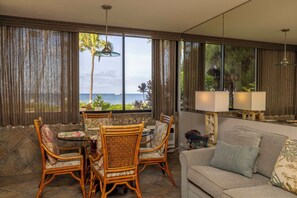 A room with a dining area, a sofa, and large windows with a view of the sea.