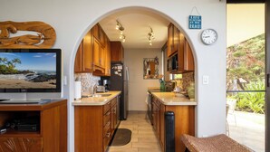 A compact kitchen with wooden cabinets, a built-in microwave, and a large TV on the left. - Oceanfront Kealia 101 Kihei Condo  Dramatic Views Steps from Pool and Beach (Kihei)