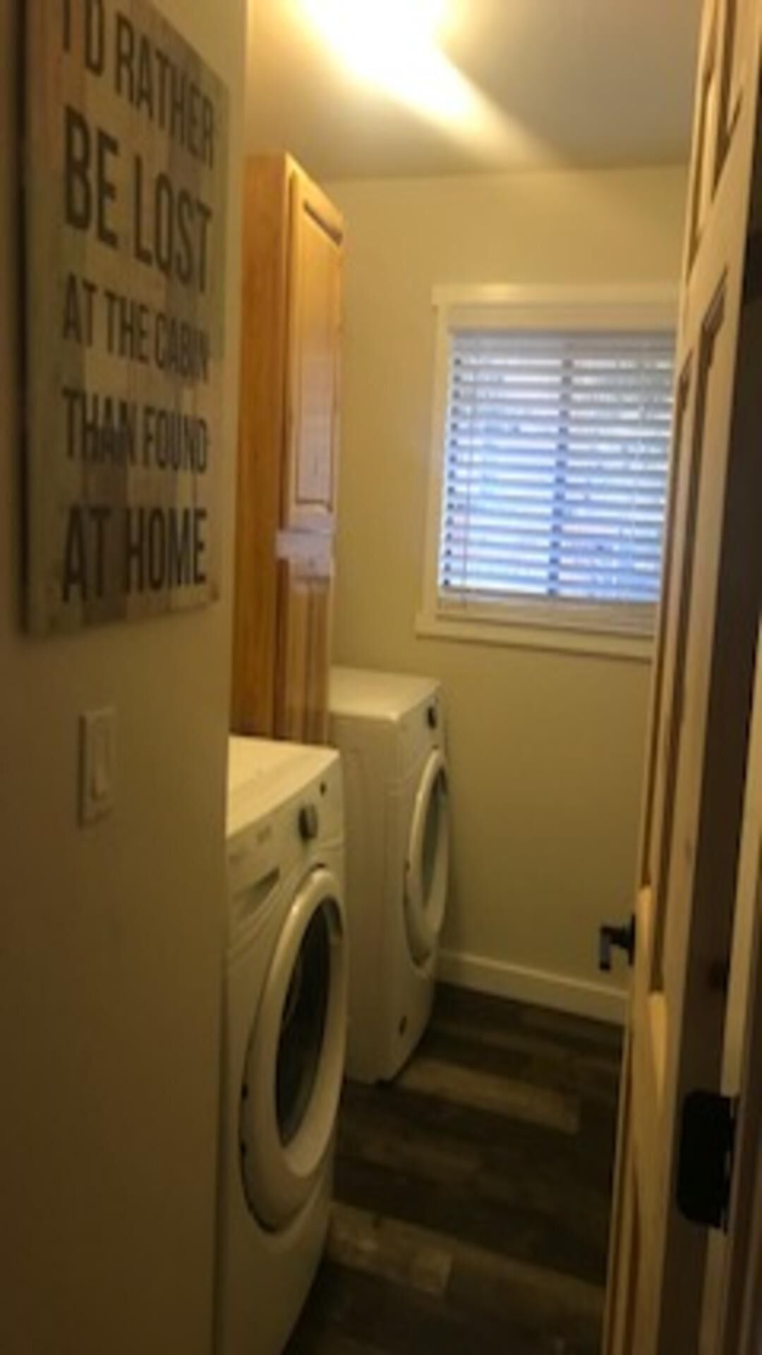A laundry room with two washing machines, a wooden floor, and a window with blinds.