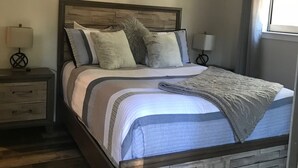 A bedroom with a bed, a ceiling fan, a bedside table with a lamp, a framed 'blessed' sign, and a window with blinds.
