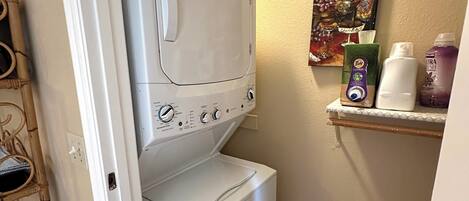 Un lavadero con lavadora y secadora de carga frontal, un estante con productos de limpieza y un cuadro en la pared.