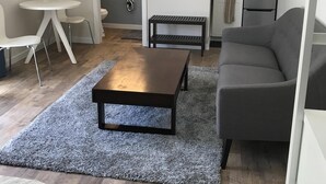 A compact living space with a grey sofa, a small coffee table, a white dining table with chairs, a kitchenette, and a bathroom with a mirror and lights. - Furnished detached garden cottage (SF)