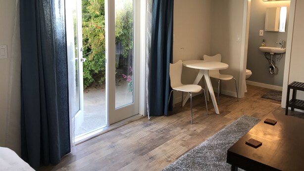 A room with a sliding glass door leading to a garden, a small round table with two chairs, a white sink, and a mirror with lights. - Furnished detached garden cottage (SF)