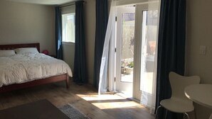 A bedroom with a bed, a small table, and a chair. There is a sliding glass door leading to an outdoor area with potted plants. - Furnished detached garden cottage (SF)