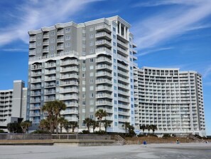 A multi-story hotel with balconies, a beach in the foreground, and a clear sky.