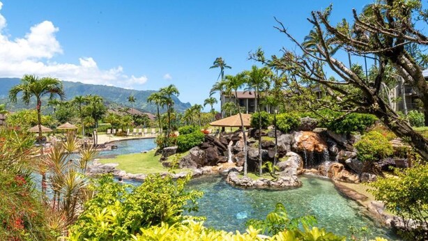 A resort with a clear pool, palm trees, and a mountainous backdrop.