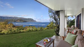 A patio with a view of the ocean and mountains, a wooden table with a plate and drinks, and a sofa with cushions.
