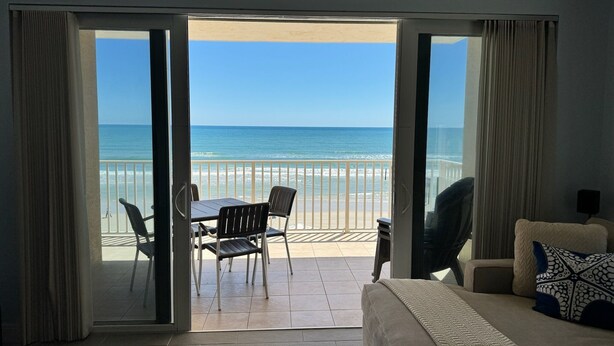 A room with a balcony overlooking a beach, a sofa, and a table with chairs.
