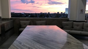 A spacious terrace with a marble table and sofas, overlooking a cityscape at sunset. - Emerald By the Sea #813, Beautiful Gulf View, Luxury Hi Rise 2 Bedroom Condo (Galveston)