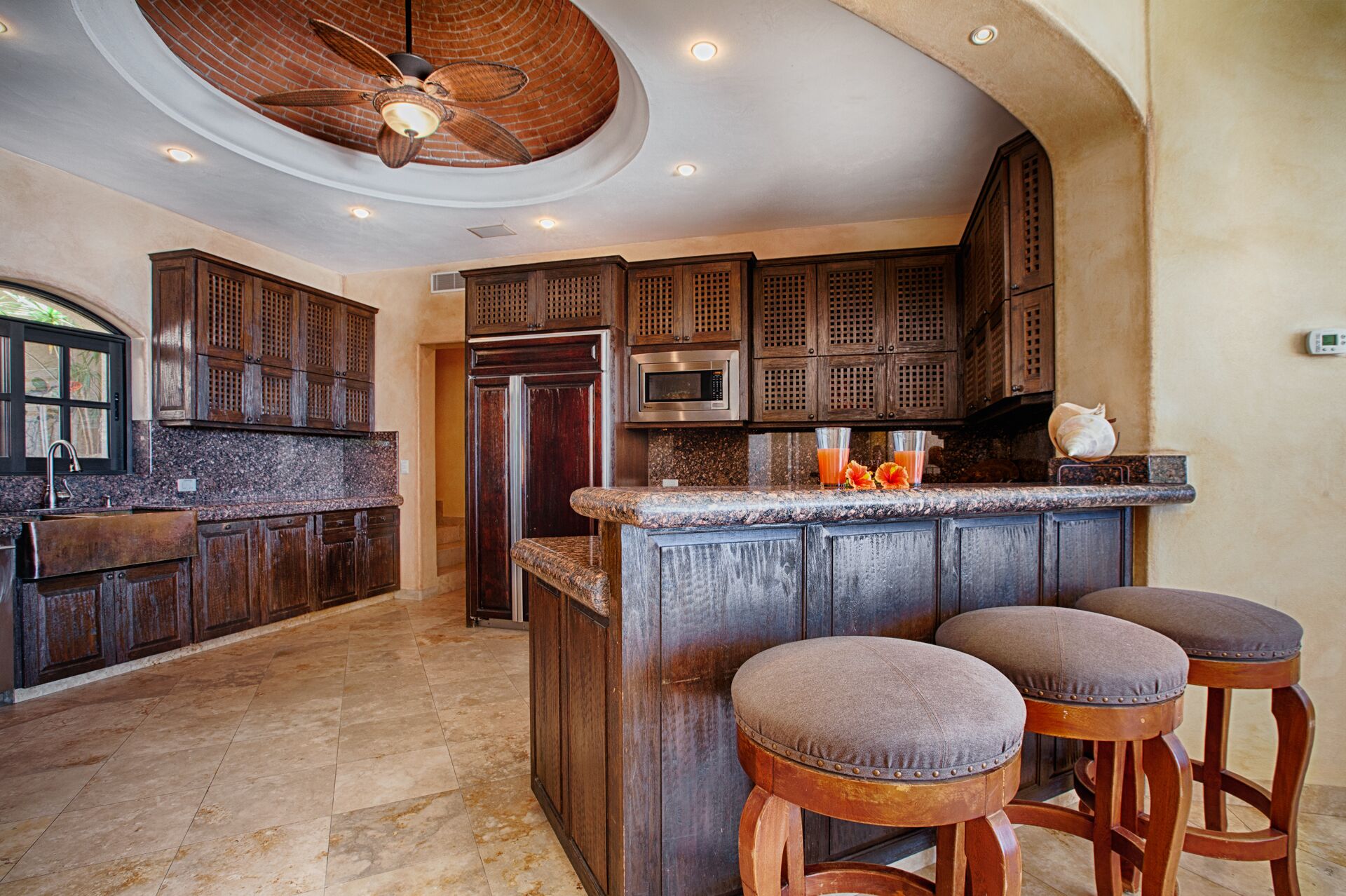 A kitchen with dark wooden cabinets, a granite countertop, a built-in microwave, and a ceiling fan. There are three wooden stools at the island.