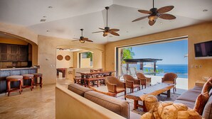 A spacious living area with a large window overlooking the ocean, a dining area with wicker furniture, and a bar with stools. - Villa Gran Vista - Gorgeous Panoramic Ocean Views Villa (Cabo San Lucas)