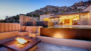 A modern outdoor pool area with a fire feature, lounge chairs, and a well-lit interior. - Villa Gran Vista - Gorgeous Panoramic Ocean Views Villa (Cabo San Lucas)