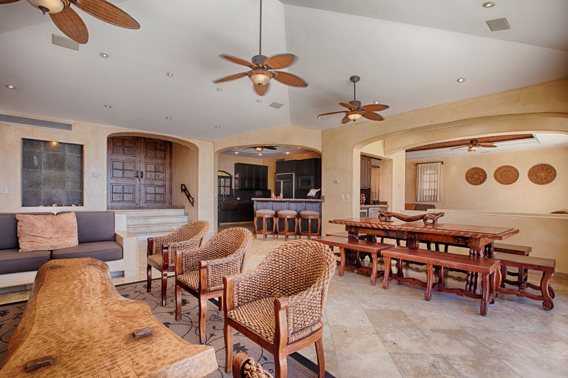 A spacious living area with a large wooden dining table, wicker chairs, a sofa, and ceiling fans.