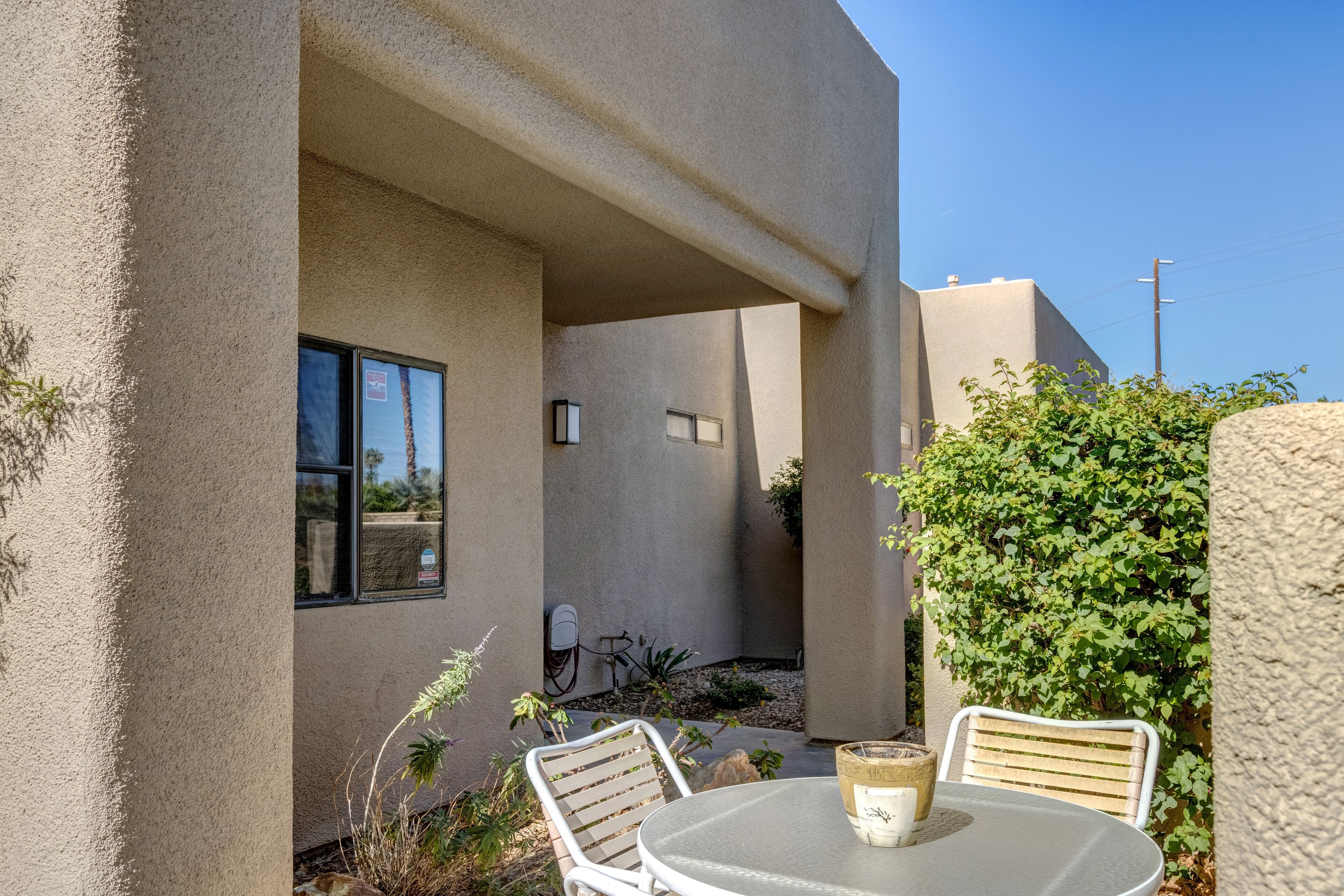 A patio with a round table and two chairs, a small garden area with plants, and a building with a stucco exterior.