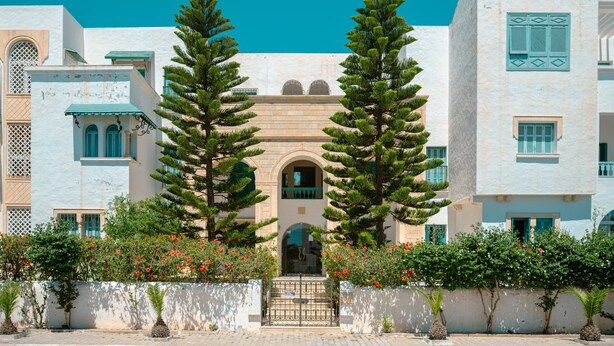 A building with a central arched entrance, flanked by two tall trees, and surrounded by a low wall with flowering plants. - Beautiful view apartment located at Yasmine Hammamet with garden, swimming pool  (Yasmine hammamet)