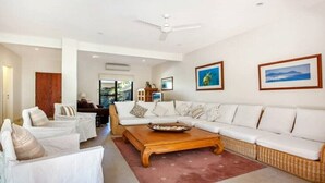 Amplia sala de estar con un sofá blanco grande, una mesa de centro de madera y sillones blancos. Se observan cuadros en las paredes y un ventilador de techo.