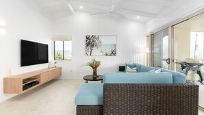 A modern living room with a flat-screen TV, a blue sofa, a wicker ottoman, a small round table with a vase, and a large window offering a view of the outdoors.