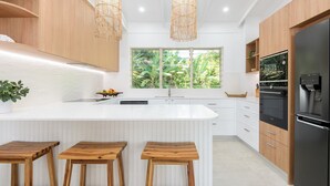 A modern kitchen with a large island, wooden stools, a refrigerator, and a window with a view of greenery.