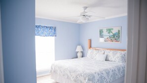 A bedroom with a bed, a ceiling fan, a window with a blue floral curtain, and a beach-themed painting on the wall. - Perfect Gulf Front Beach House *Beach Club Amenities*The Best of Both Worlds! (Gulf Shores)