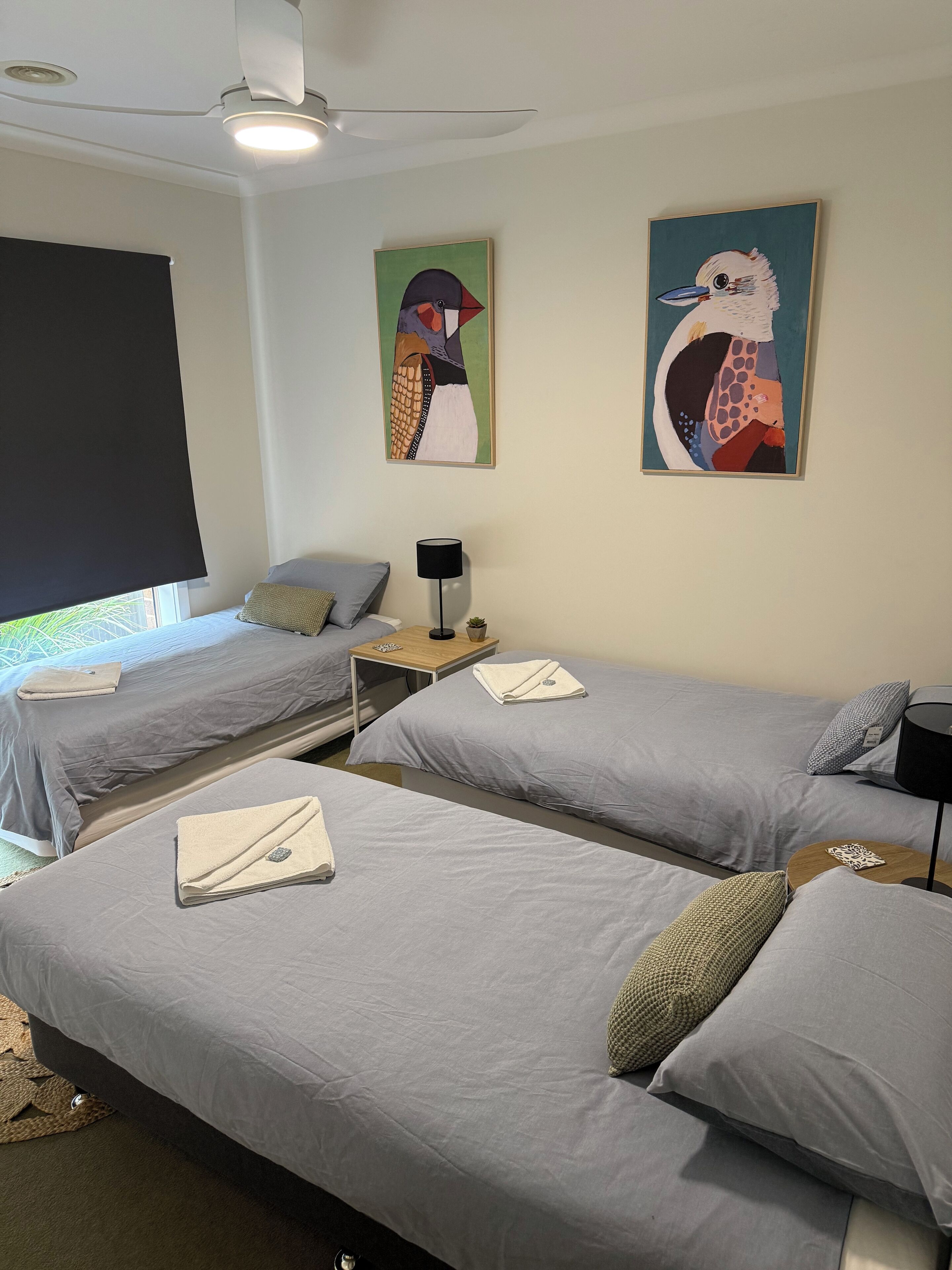 A hotel room with two single beds, a ceiling fan, artwork on the walls, a small table with a lamp, and a window with a view.