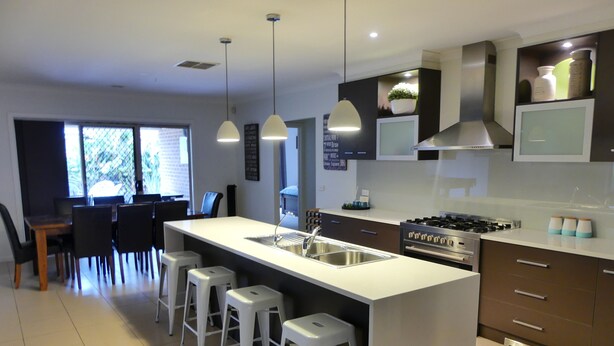 A modern kitchen with a central island, bar stools, a stove, and a dining area with chairs and a table. - Parkins Fairway - Golf, Lake & Relaxation (Yarrawonga)