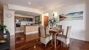 A modern kitchen with wooden cabinets and a central island, a dining area with wicker chairs and a wooden table, and a painting of a beach scene on the wall. - 5 The Point Apartments  |  Port Douglas (PORT DOUGLAS)