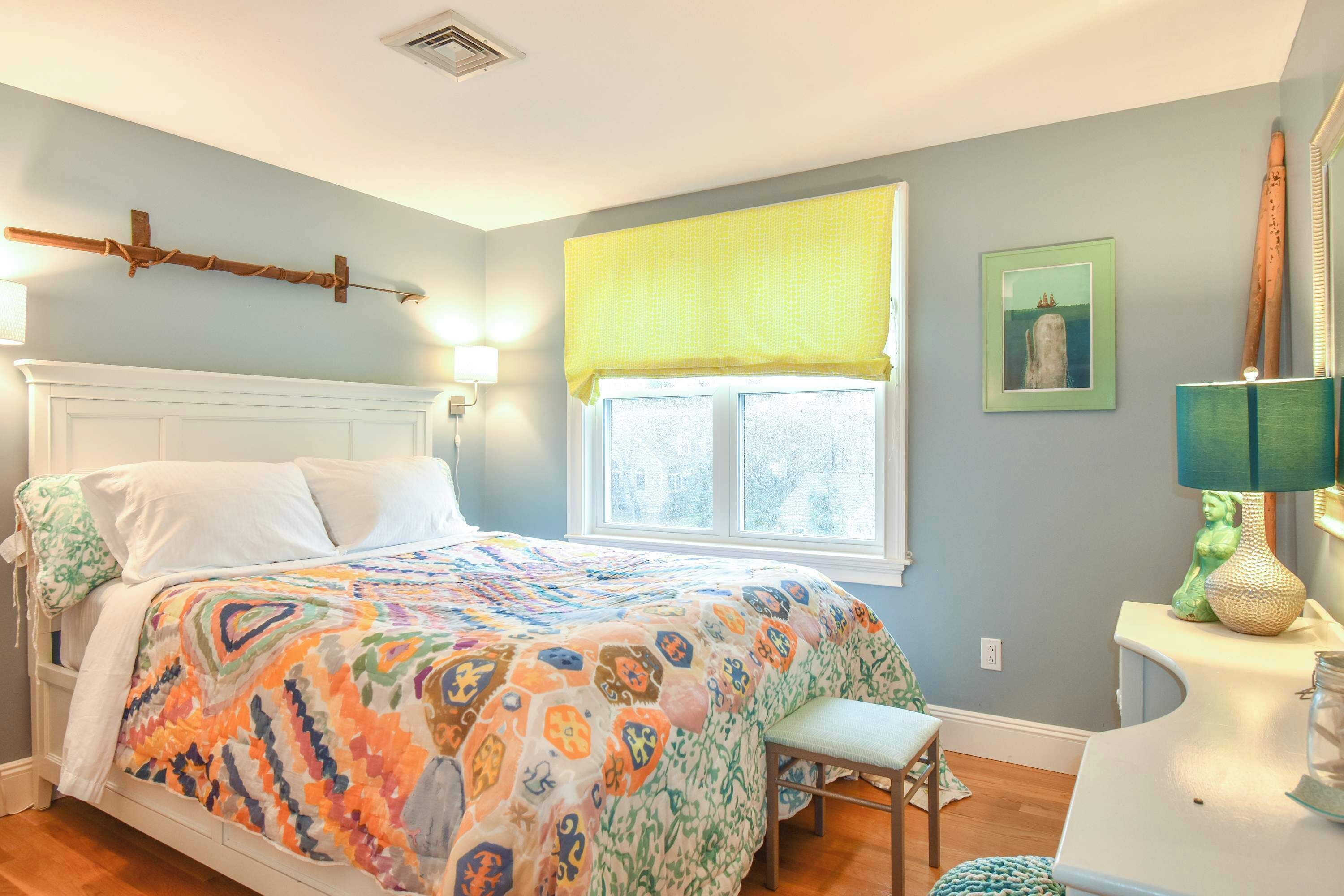 Un dormitorio con cama, ventana con cortinas amarillas, lámpara de noche y un cuadro en la pared.