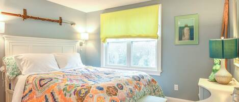 Un dormitorio con cama, ventana con cortinas amarillas, lámpara de noche y un cuadro en la pared.