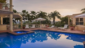 Zona de piscina con sillones de descanso y sombrillas, rodeada de palmeras y vistas al océano.