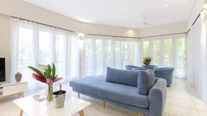 Una sala moderna con un sofá, una mesa de centro y un televisor. Hay ventanas amplias con cortinas translúcidas, un ventilador de techo y una planta en el alféizar.