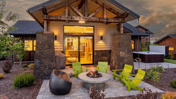 A rustic cabin with a stone facade, a wooden deck with green chairs, and a fire pit. - Plush beds ~ heated resort pool ~ hot tub ~ Tetherow/Bend/Bachelor ~ wifi ~ 🐶 (Bend)