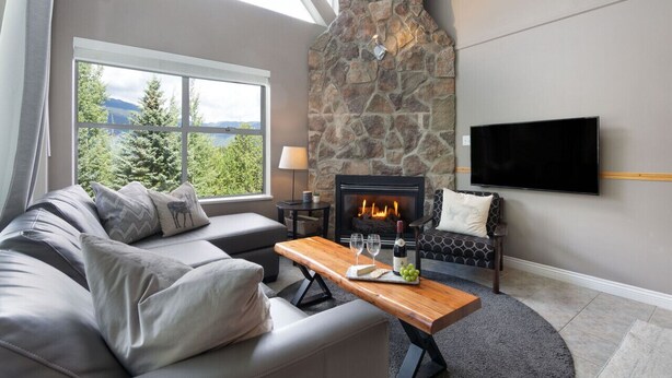 A living room with a stone fireplace, a sofa, a coffee table with wine glasses and a bottle, and a TV mounted on the wall. - Mountain-Side 3Br/2Ba - HotTub/Views/Free Parking (Whistler)