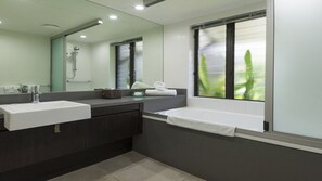 A modern bathroom with two sinks, a bathtub, a large mirror, and a window with a view of greenery.