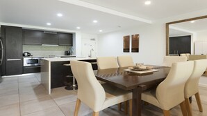 A modern kitchen with dark cabinets, a central island, and a dining area with a wooden table and beige chairs. - Azure 125-126 | Port Douglas (PORT DOUGLAS)