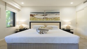 A hotel room with a large bed, two bedside tables with lamps, a painting above the bed, and a window on the left. - Azure 125-126 | Port Douglas (PORT DOUGLAS)