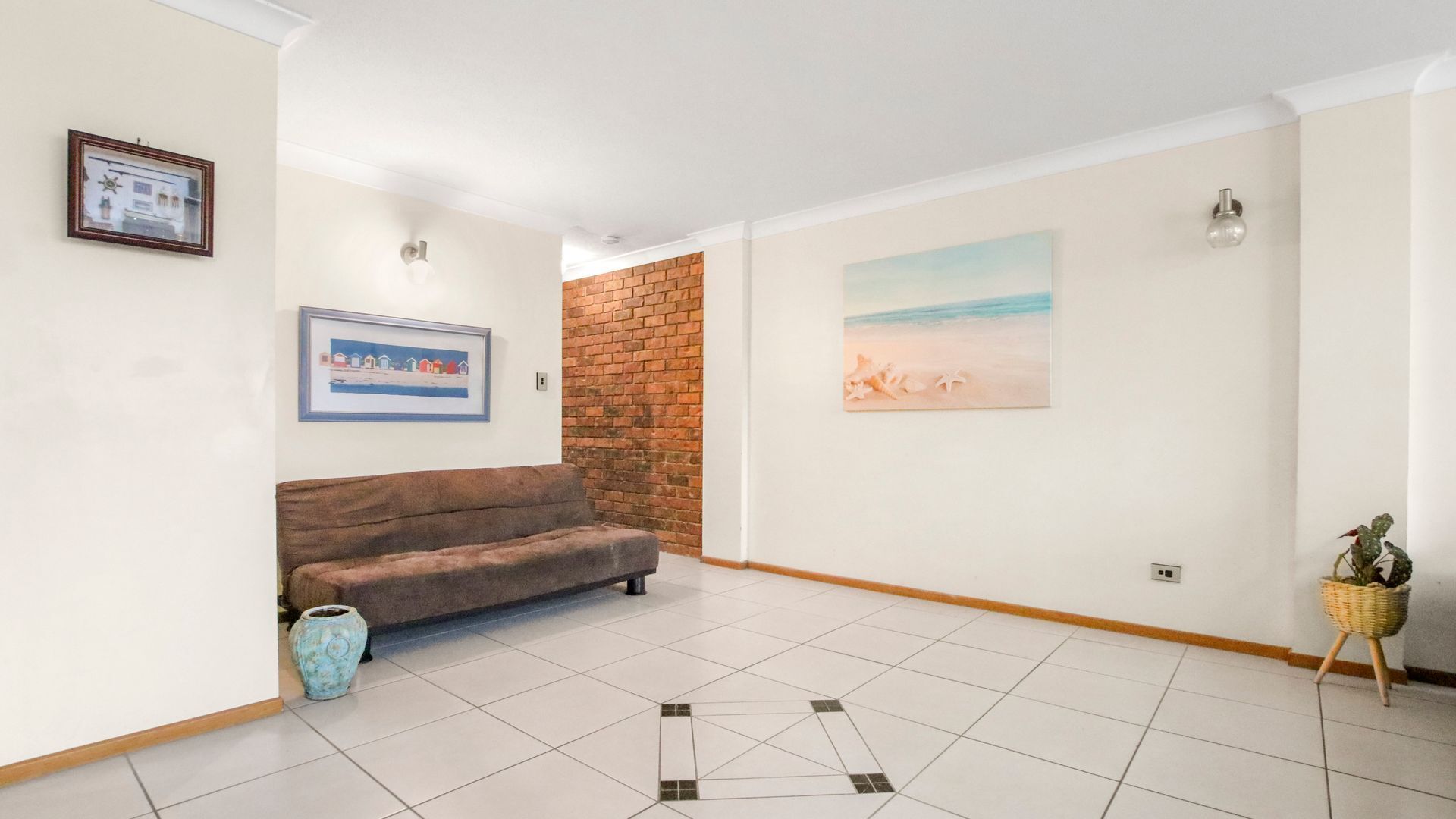 A spacious room with a brown sofa, a tiled floor, a brick wall, and two framed artworks.