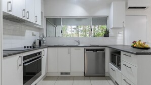A modern kitchen with white cabinetry, a stainless steel dishwasher, and a window with a view of greenery. - 99 Reef Resort  |  Port Douglas (PORT DOUGLAS)