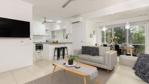 A modern apartment with a kitchen, dining area, and living room. It includes a flat-screen TV, ceiling fan, and large windows. - 99 Reef Resort  |  Port Douglas (PORT DOUGLAS)